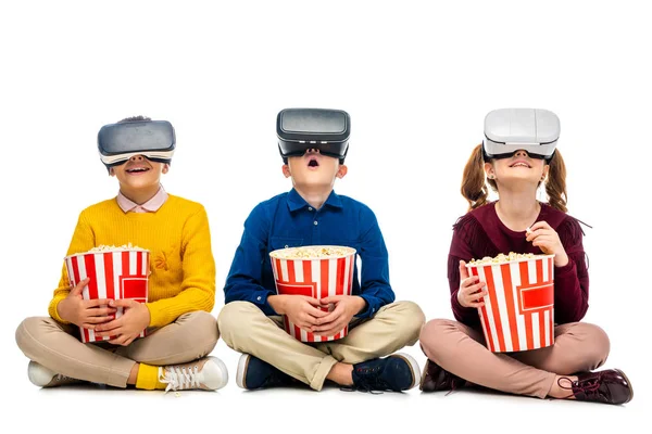 Amazed children with virtual reality headsets on heads holding striped buckets and eating popcorn isolated on white — Stock Photo