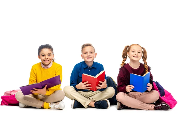 Escolares alegres sentados, sosteniendo libros y mirando a la cámara aislada en blanco - foto de stock