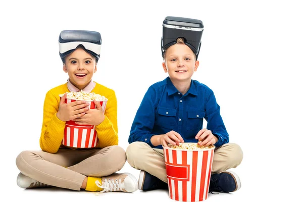 Aluna surpreso com fones de ouvido realidade virtual na cabeça segurando balde de papelão listrado e menino comendo pipoca isolada no branco — Fotografia de Stock