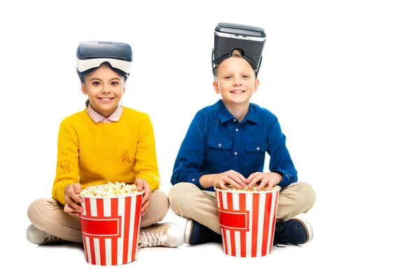 Niños sonrientes con auriculares de realidad virtual en cabezas que sostienen cubos de palomitas de maíz a rayas y mirando a la cámara aislada en blanco - foto de stock