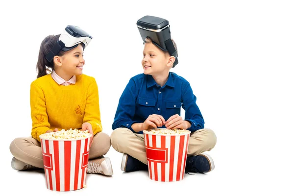 Lächelnde Kinder mit Virtual-Reality-Headsets auf Köpfen, die neben gestreiften Popcorn-Eimern sitzen und einander isoliert auf weißem Grund anschauen — Stockfoto