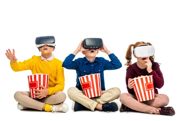 Verblüffte Kinder mit Virtual-Reality-Headsets auf Köpfen, die gestreifte Eimer halten und Popcorn isoliert auf weiß essen — Stockfoto