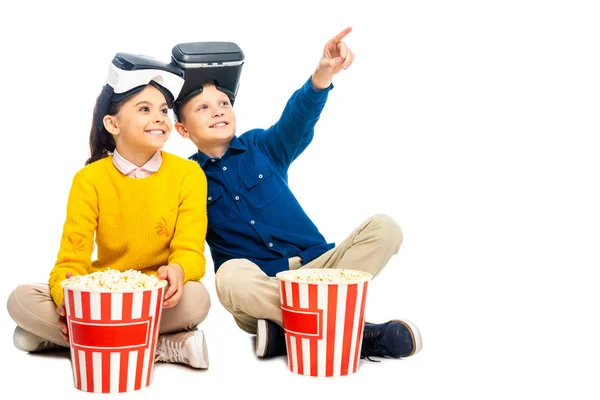 Smiling boy with virtual reality headsets on head pointing with finger and cute schoolgirl holding striped popcorn bucket isolated on white — Stock Photo