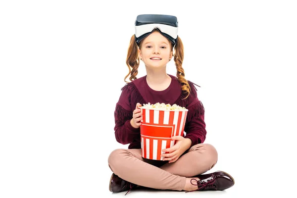 Niño con auriculares de realidad virtual en la cabeza sosteniendo palomitas de maíz a rayas cubo y mirando a la cámara aislada en blanco - foto de stock