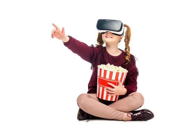 Kind mit Virtual-Reality-Headset auf dem Kopf hält gestreiften Popcorn-Eimer und zeigt mit dem Finger isoliert auf weiß — Stockfoto