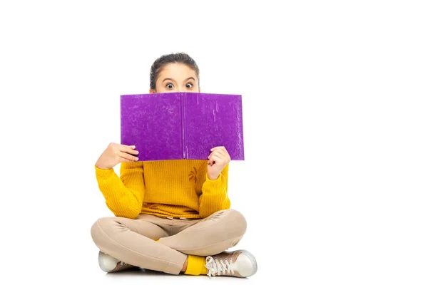 Colegiala en suéter amarillo sentado y mirando por encima del libro púrpura en la cámara aislada en blanco - foto de stock