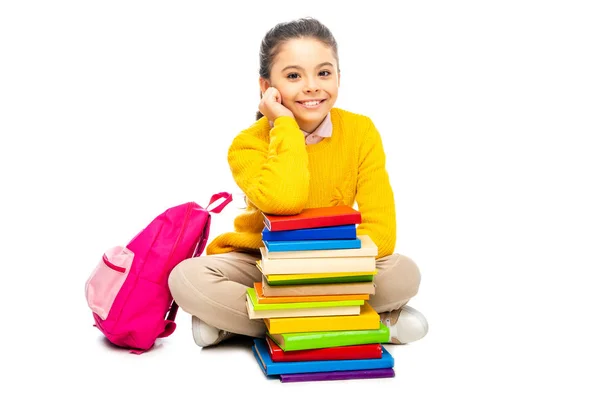 Cute schoolgirl sitting near stack of books and pink backpack and looking at camera isolated on white — Stock Photo