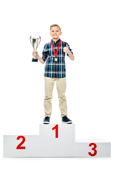 Sorrindo menino de pé no pedestal vencedor, segurando copo do troféu, mostrando o polegar para cima e olhando para a câmera isolada no branco — Fotografia de Stock