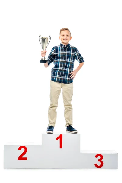 Menino feliz de pé no pódio vencedor, segurando copo do troféu, sorrindo e olhando para a câmera isolada no branco — Fotografia de Stock