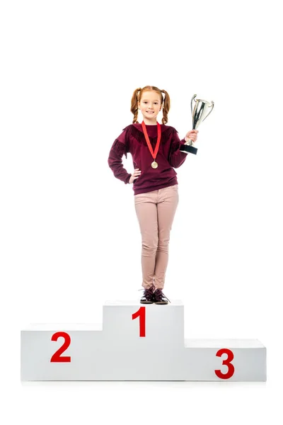 Smiling schoolgirl with medal standing on winner podium, holding trophy cup and looking at camera isolated on white — Stock Photo