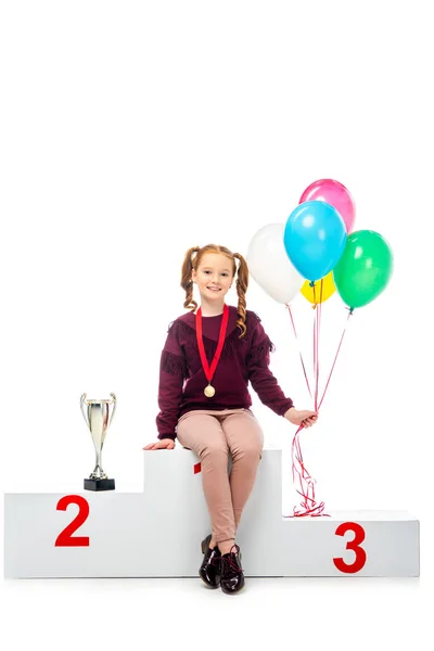 Smiling schoolgirl sitting on winner podium near trophy cup, holding colorful balloons and looking at camera isolated on white — Stock Photo