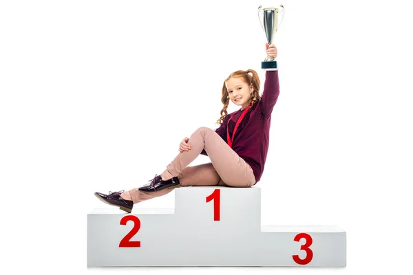 Smiling schoolgirl sitting on winner podium, holding trophy cup in raised hand and looking at camera isolated on white — Stock Photo