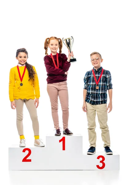 Smiling preteen kids with medals and trophy cup standing on winner pedestal, smiling and looking at camera isolated on white — Stock Photo