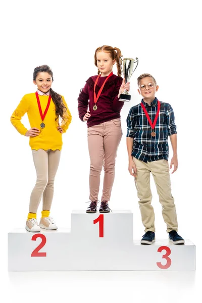 Crianças felizes com medalhas e taça de troféu em pé no pedestal vencedor, sorrindo e olhando para a câmera isolada no branco — Fotografia de Stock