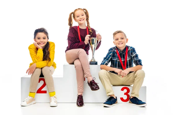 Bambini felici con medaglie e coppa trofeo sorridente, seduti sul piedistallo vincitore e guardando la fotocamera isolata sul bianco — Foto stock