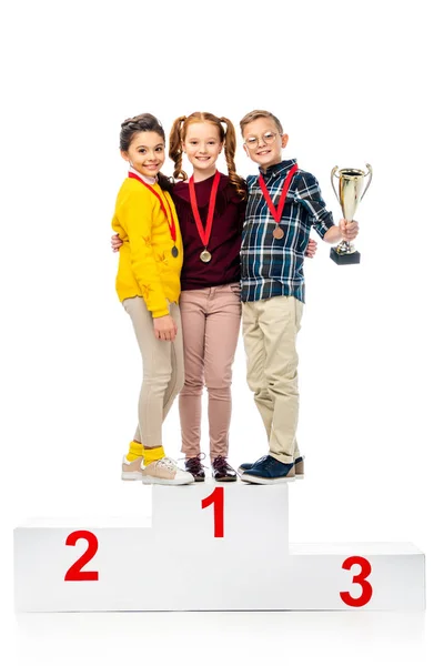 Niños felices con medallas y copa de trofeo sonriendo y de pie en el podio ganador y mirando a la cámara aislada en blanco - foto de stock