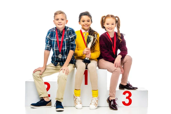 Bambini sorridenti con medaglie e coppa trofeo seduti sul podio del vincitore e guardando la fotocamera isolata sul bianco — Foto stock