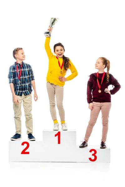 Schoolchildren with medals standing on winner podium and looking at smiling schoolgirl holding trophy cup over head isolated on white — Stock Photo