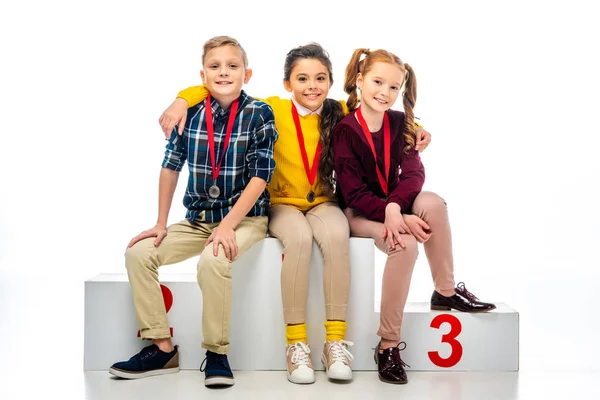 Bambini sorridenti con medaglie seduti sul podio vincitore e guardando la fotocamera isolata sul bianco — Foto stock