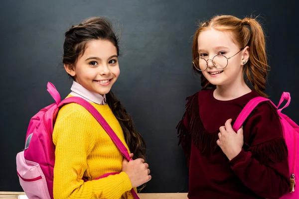 Fröhliche Schulmädchen mit rosa Rucksäcken, die in die Kamera schauen und auf schwarzem Hintergrund lächeln — Stockfoto