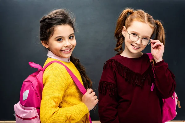 Lächelnde Schulmädchen mit rosa Rucksäcken schauen in die Kamera auf schwarzem Hintergrund — Stockfoto