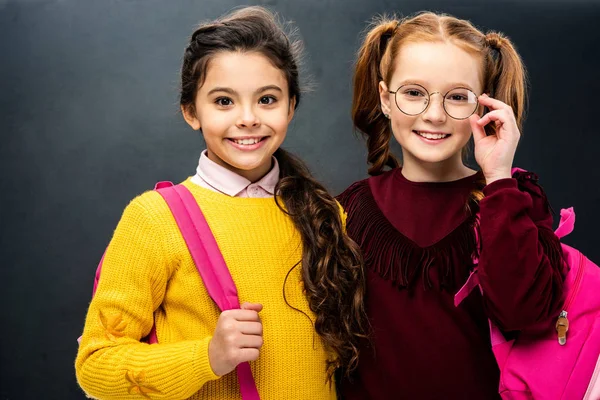 Cute schoolgirls with backpacks smiling and looking at camera on black background — Stock Photo