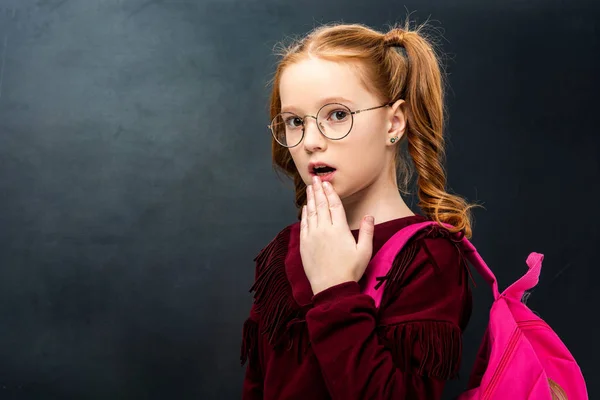 Surprised schoolgirl in glasses with pink backpack looking at camera on black background — Stock Photo