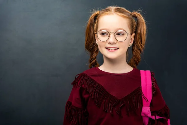 Cute schoolgirl in glasses with pink backpack looking at camera on black background — Stock Photo
