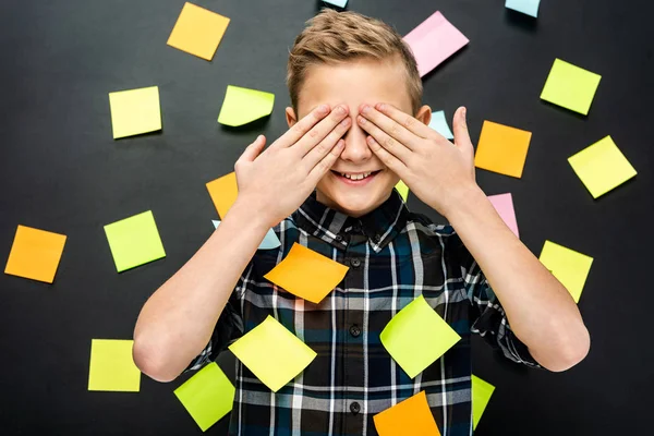 Smiling boy with multicolored stickers covering eyes with hands on black background — Stock Photo