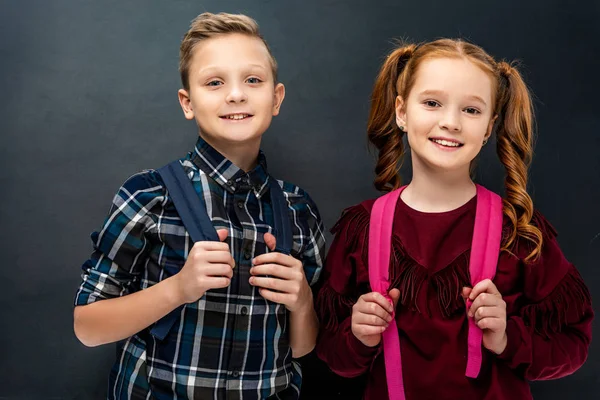 Smiling schoolchildren with backpacks looking at camera on black background — Stock Photo