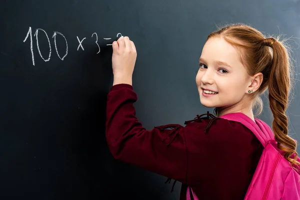 Schoolgirl with pink backpack writing on blackboard with chalk and looking at camera — Stock Photo