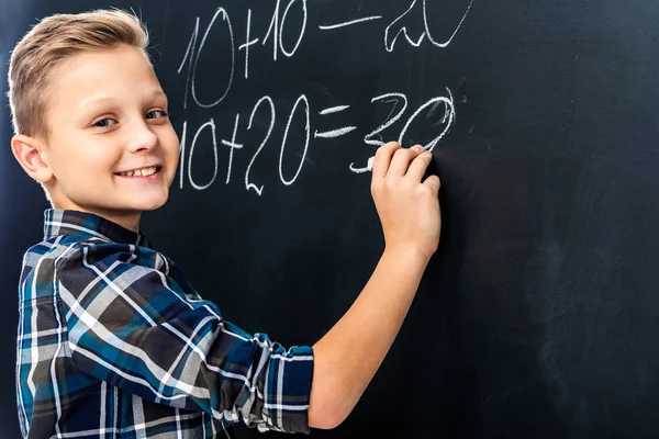 Niño sonriente escribiendo ejemplo de matemáticas en pizarra con tiza y mirando a la cámara - foto de stock