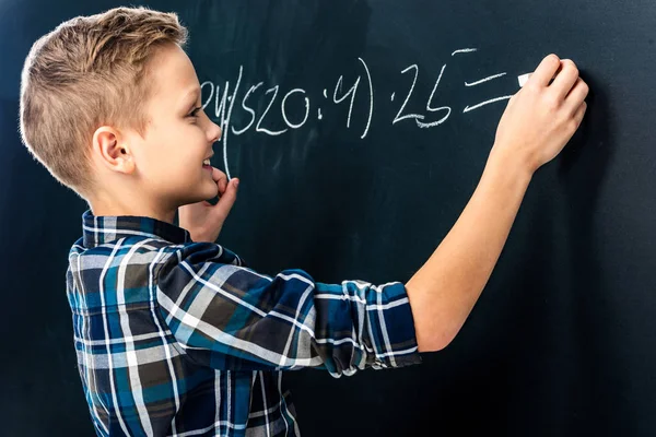Niño sonriente escribiendo ejemplo de matemáticas en pizarra con tiza - foto de stock