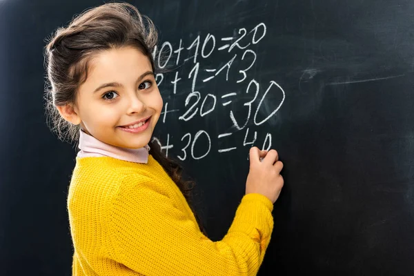 Smiling schoolgirl writing on blackboard with chalk and looking at camera — Stock Photo