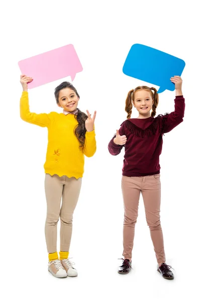 Colegialas alegres sosteniendo burbujas de habla multicolor sobre la cabeza, mostrando el pulgar hacia arriba y signo de paz y mirando a la cámara aislada en blanco - foto de stock