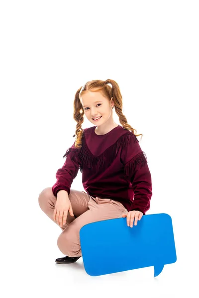 Colegiala sonriente sentada en hunkers, sosteniendo la burbuja del habla y mirando a la cámara aislada en blanco - foto de stock