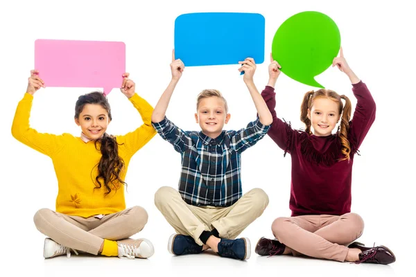 Smiling kids sitting and holding speech bubbles over heads isolated on white — Stock Photo