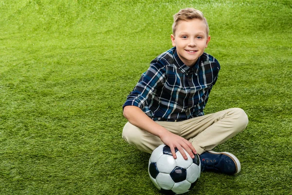 Niño sonriente sentado en el césped con la mano en la pelota de fútbol y mirando a la cámara - foto de stock