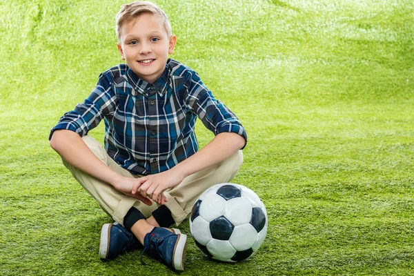 Garçon souriant assis sur la pelouse près du ballon de football et regardant la caméra — Photo de stock