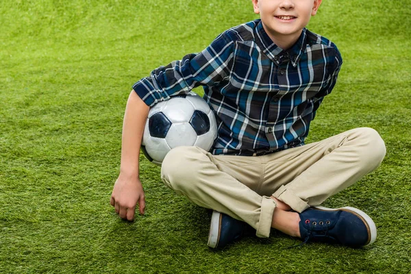 Vista cortada de menino sorridente segurando bola de futebol e sentado no gramado — Fotografia de Stock