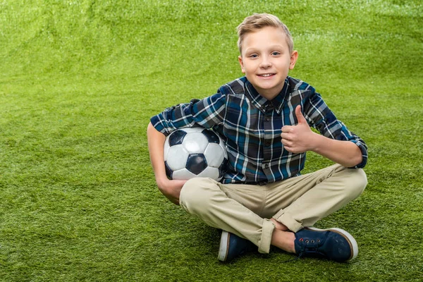 Garçon souriant tenant ballon de football, regardant la caméra et montrant pouce sur la pelouse — Photo de stock