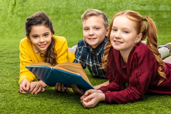 Cute preteen schoolkids lying on green lawn, holding book and looking at camera — Stock Photo