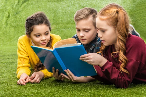 Милые школьники, лежащие на зеленой лужайке и читающие книгу — стоковое фото