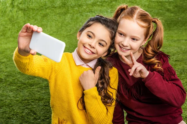 Smiling schoolgirls taking selfie and showing thumb up and peace sign on lawn — Stock Photo