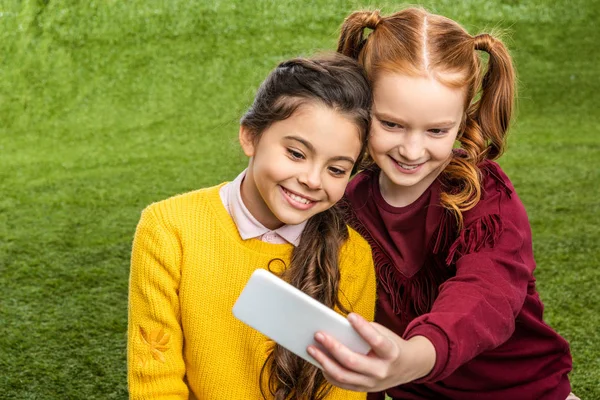 Cute schoolgirls smiling and taking selfie on lawn — Stock Photo