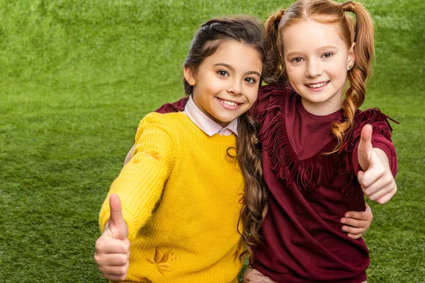 Smiling schoolgirls looking at camera and showing thumbs up on lawn — Stock Photo