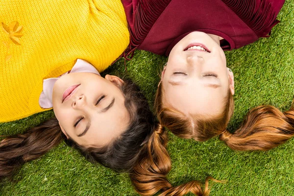 Vue de dessus des écolières mignonnes avec les yeux fermés couché tête à tête sur la pelouse — Photo de stock