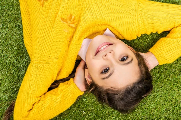 Вид сверху на улыбающуюся школьницу в желтом свитере с руками на голове, смотрящую в камеру и лежащую на лужайке — стоковое фото