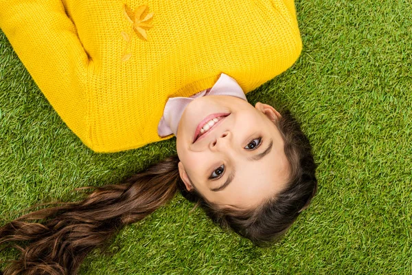 Вид зверху мила школярка в жовтому светрі з довгим волоссям, що лежить на газоні і дивиться на камеру — стокове фото