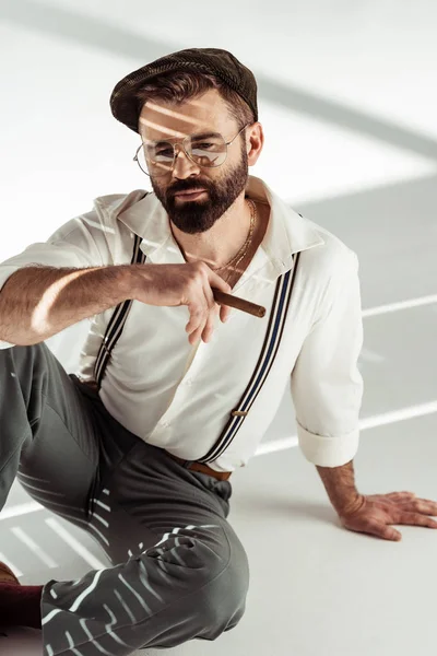 Вдумливий бородатий чоловік в окулярах і кепці сидить на підлозі і тримає сигару — стокове фото